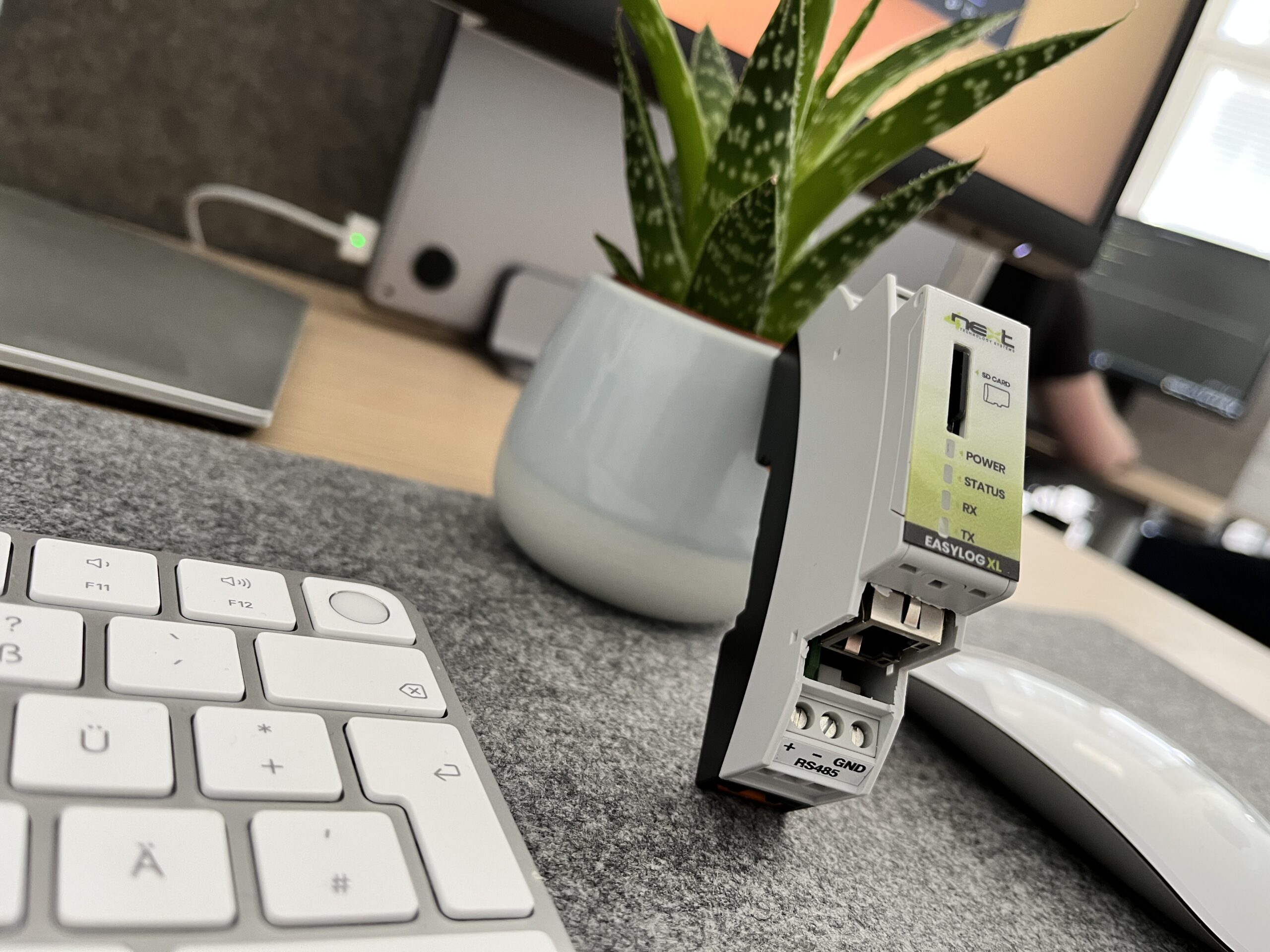 A close-up of a computer keyboard, mouse, and an ewon flexy device in front of a potted plant on a desk, with a monitor and blurred background—perfect for an easy start in IIoT.