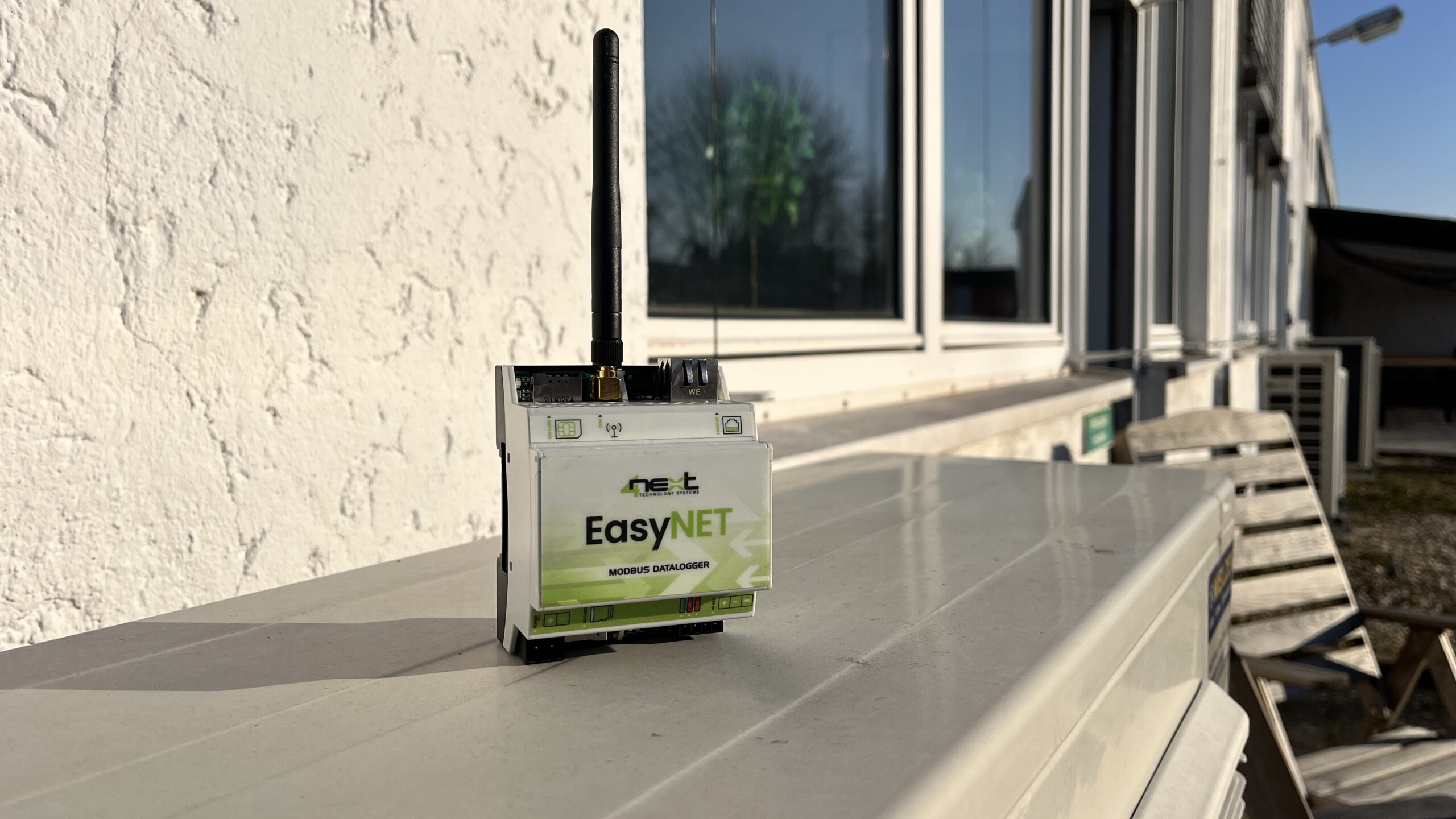 A small device labeled EasyNET with an antenna stands on top of an outdoor unit against a white stucco wall, showcasing IIoT platform capabilities, with windows reflecting sunlight in the background.