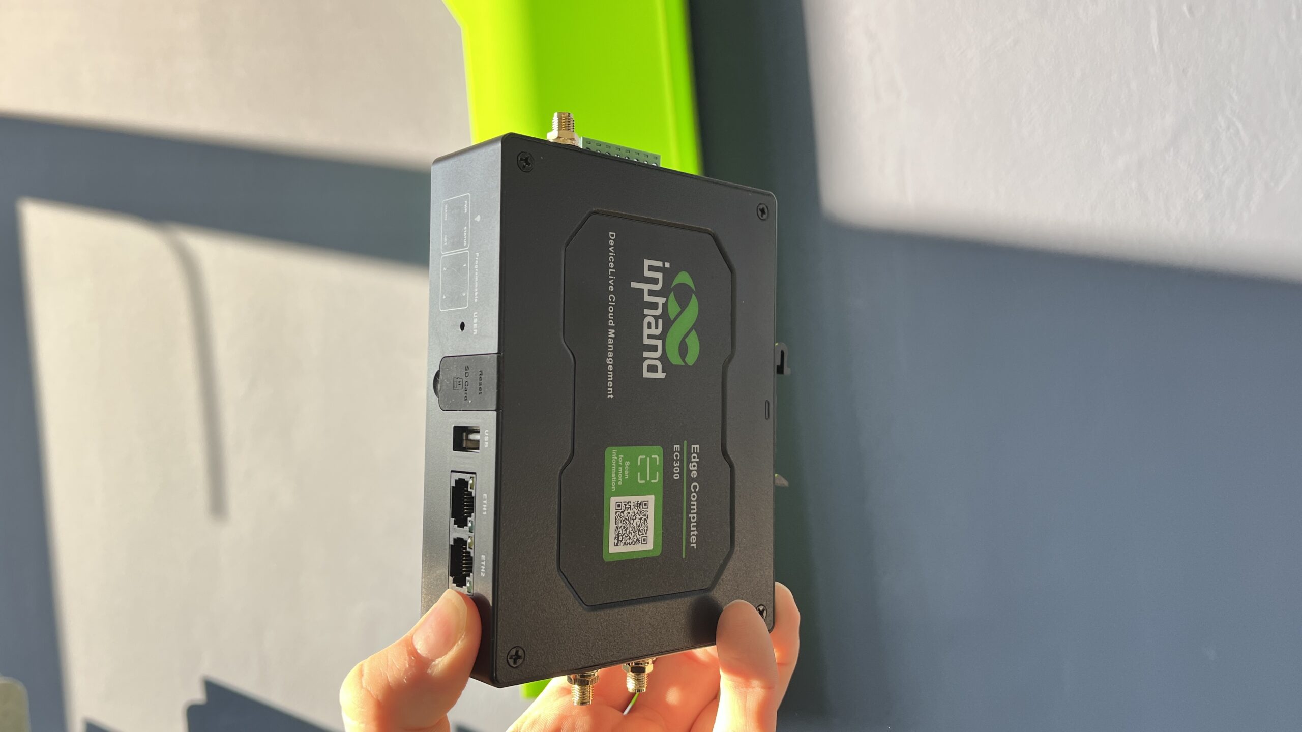 A person holds a black UPRAND device with various ports and a green label, against a wall with sunlight casting shadows—showcasing compatibility with the ewon flexy IIoT platform.