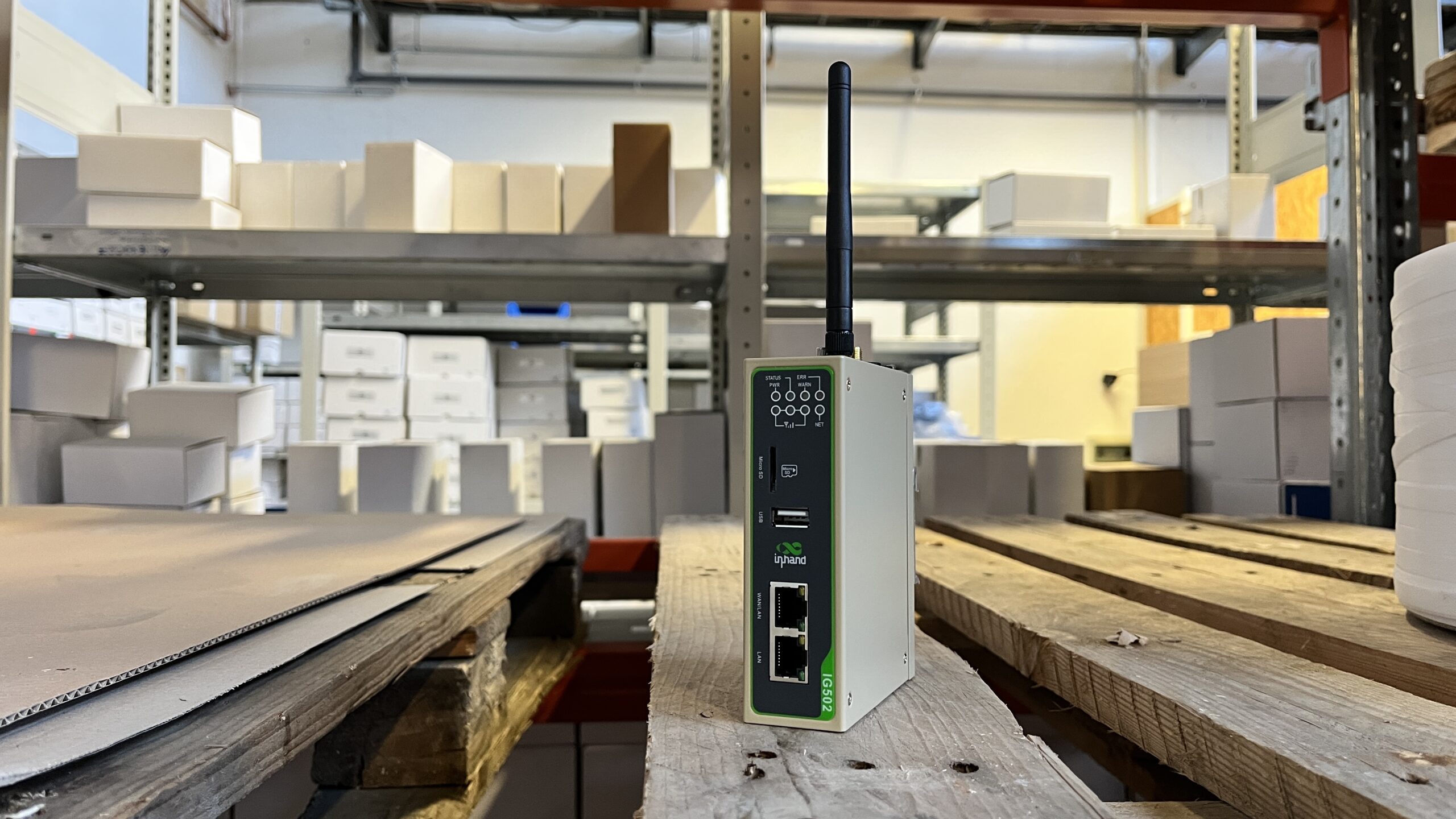A small industrial router, such as an ewon flexy, with an antenna sits on a wooden pallet in a warehouse, surrounded by shelves filled with white boxes and packages, ready to connect machines to your IIoT platform.