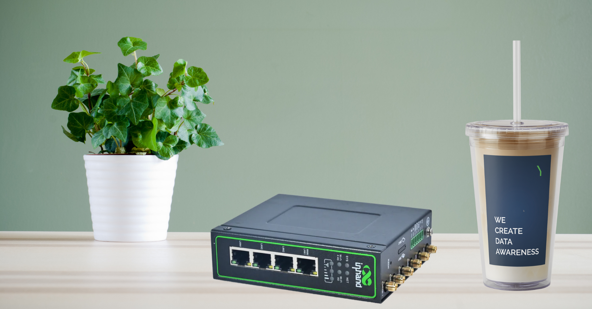 A potted green plant, a black network switch, and a plastic iced coffee cup with WE CREATE DATA AWARENESS printed on it, all placed on a light wooden surface against a green wall.