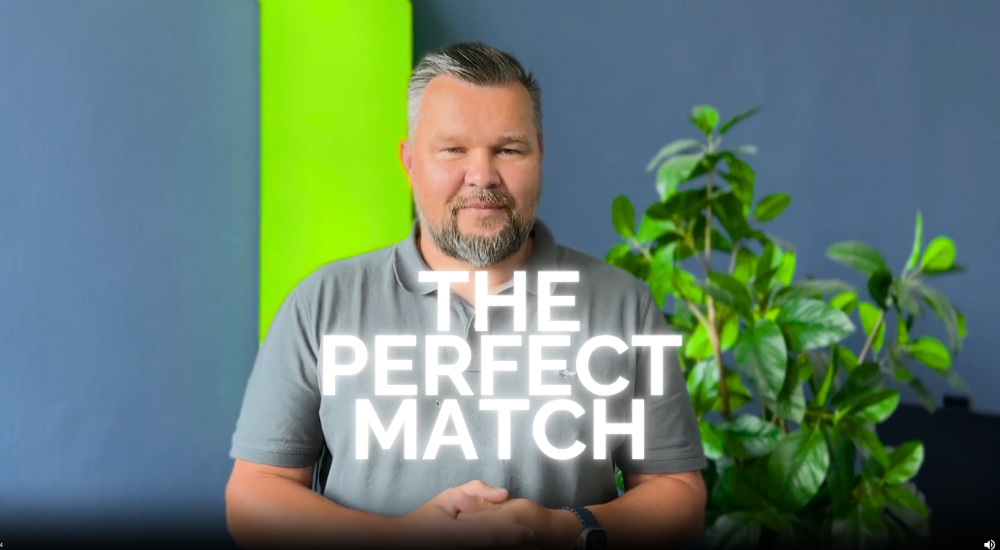 visualys- A man with a beard stands indoors in front of a green plant and a bright green and blue wall. Bold, white text across his chest reads THE PERFECT MATCH.