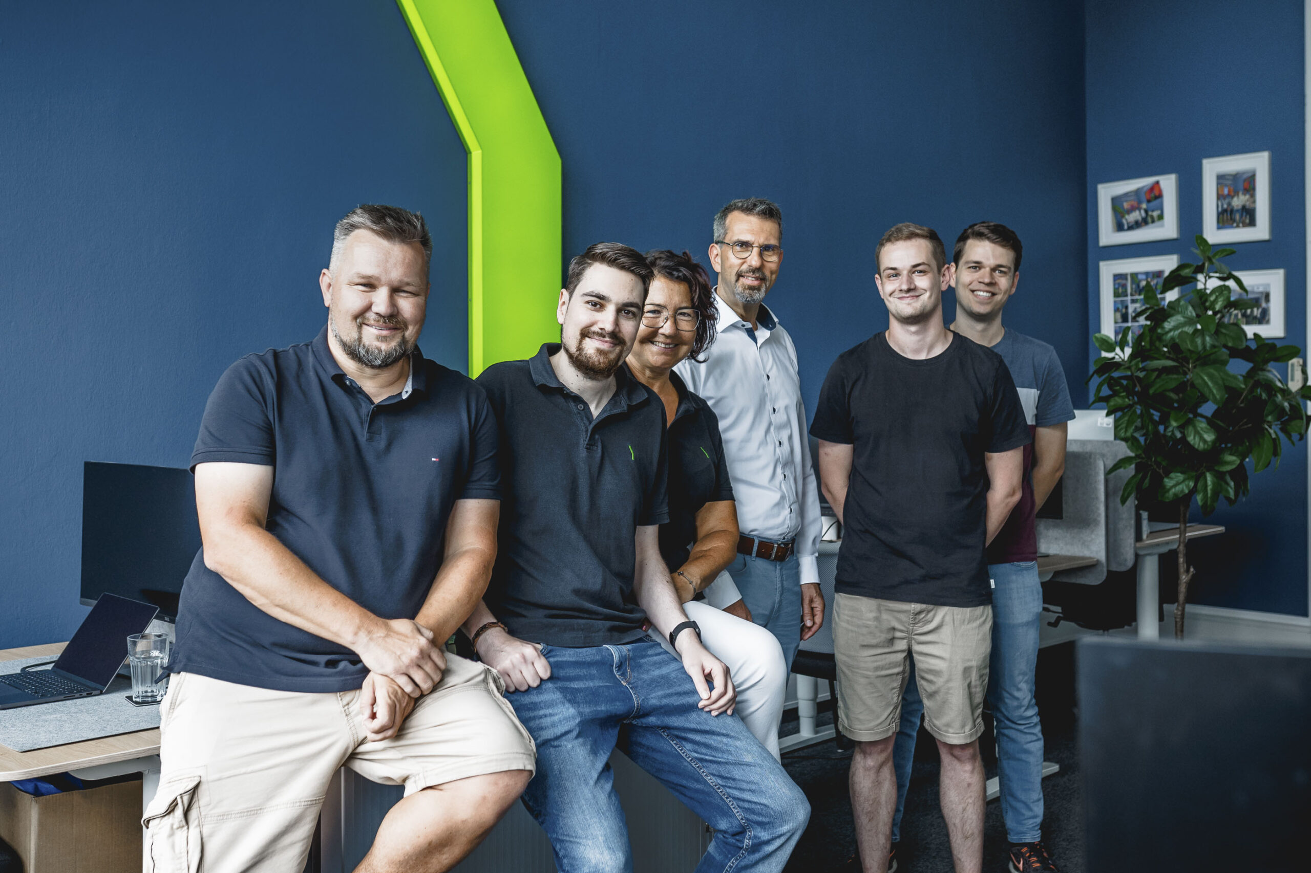 Six people, five men and one woman, stand and sit together in a modern office with blue walls, a green accent, and framed photos. They are smiling and casually dressed, creating a friendly and relaxed atmosphere.