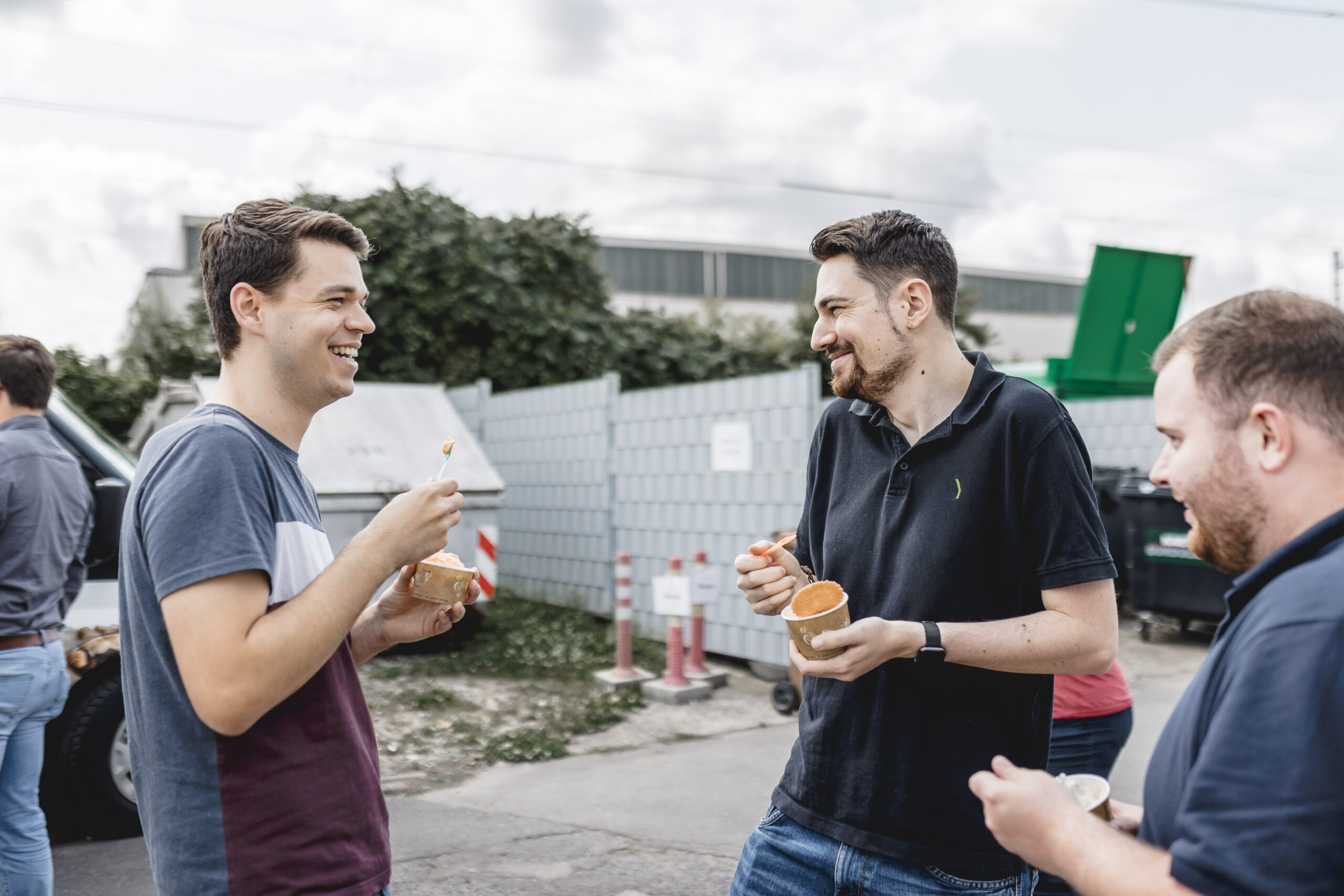 Three men stand outside laughing and eating food from small containers with forks, with a fence, greenery, and cloudy sky in the background.