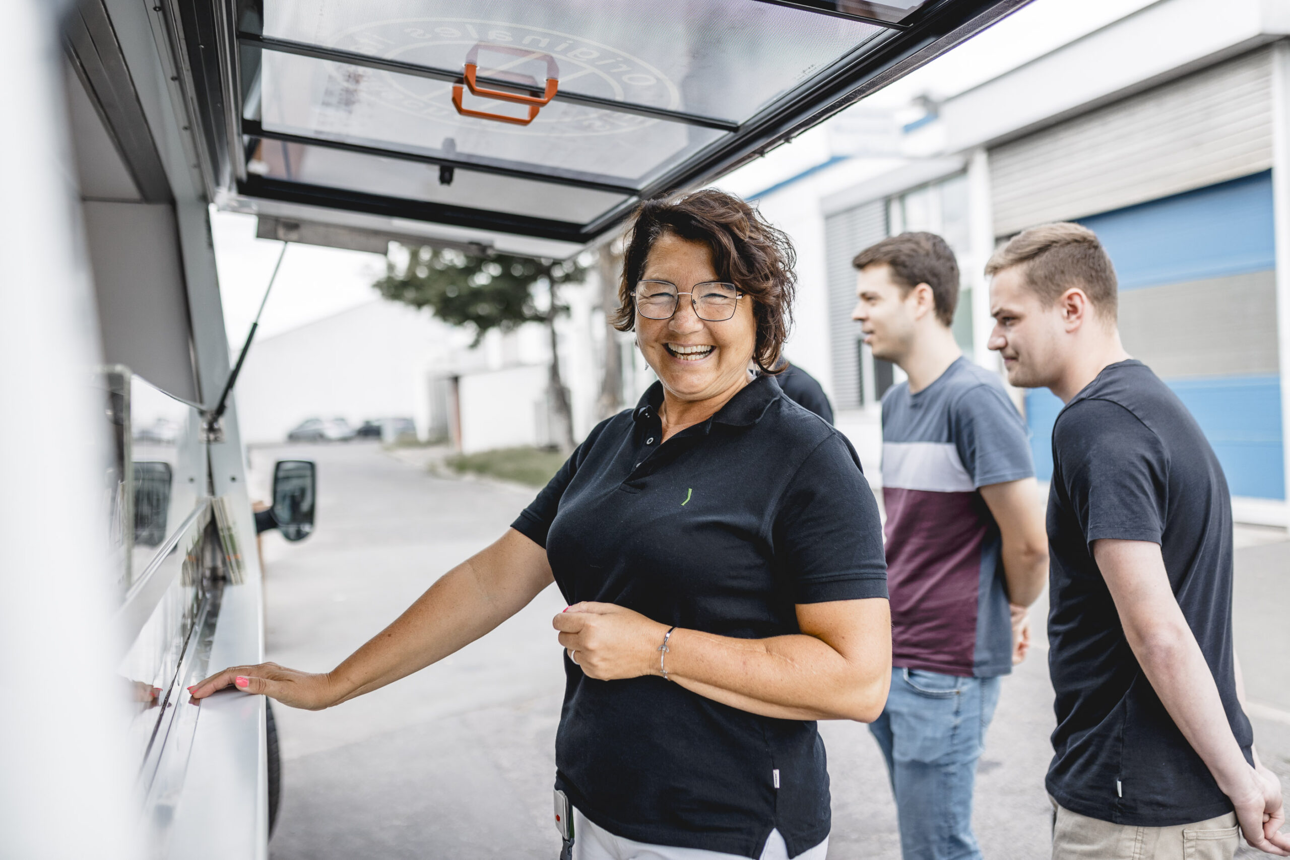 A smiling woman stands at a food truck window, holding a menu, with two men waiting in line behind her. The scene takes place outdoors near a building with blue doors.